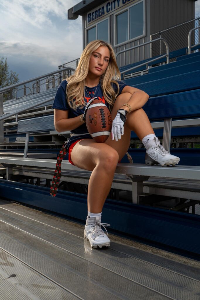 high school senior portrait of girl holding football and witting on bleachers