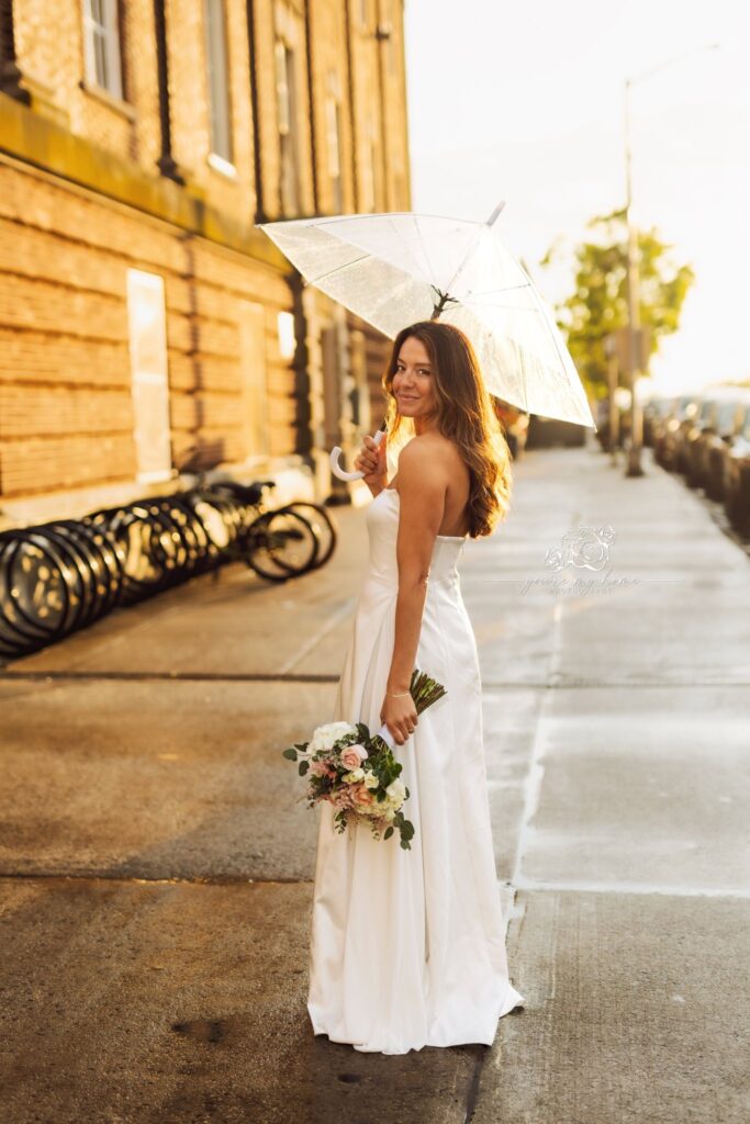 woman holding bouquet and umbrella looking over should, smiling with light behind her