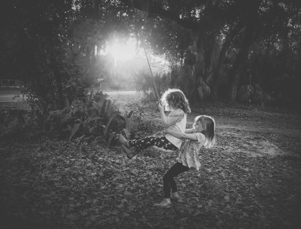 black and white photo of kids playing outside, one pushing the other on a swing